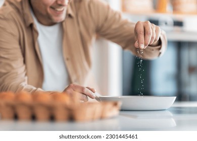 A close-up of a male's hands, salting eggs that he making himself for breakfast in the sunny kitchen.