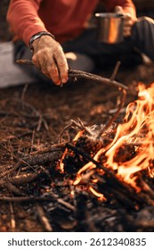 Close-up of a man adding a stick to a campfire in an outdoor setting, holding a mug. The scene suggests camping and enjoying nature with warmth and rustic simplicity.