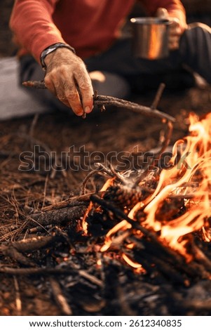 Close-up of a man adding a stick to a campfire in an outdoor setting, holding a mug. The scene suggests camping and enjoying nature with warmth and rustic simplicity.