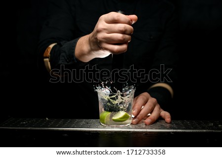 Close-up. Man bartender surely holds black madler in his hand and squeeze slices of citrus to cocktail