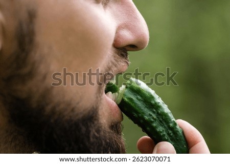 Close-up of a man with a beard taking a bite of a crisp, green cucumber. The focus is on the cucumber and the man's mouth, with a blurred green background.