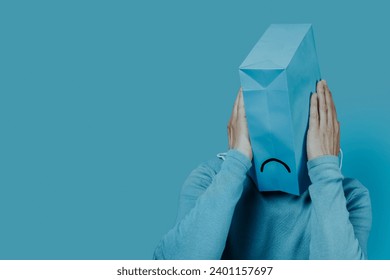 closeup of a man with a blue paper bag in his head with a sad mouth drawn in it, and his hands around it, on a blue background with some blank space on the left
