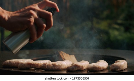 Closeup Man Chef Hands Spraying Oil On Sausages On Grill On Backyard. Unrecognized Man Cooking Sausages On Grill Outdoors. Unknown Man Preparing Bbq Sausages Outside.