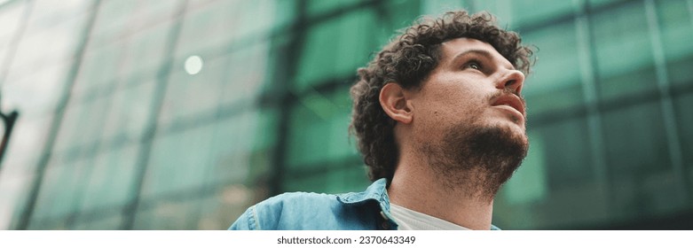 Close-up, man in denim shirt walks down the street, stops and looks around at the upper floors of modern buildings - Powered by Shutterstock