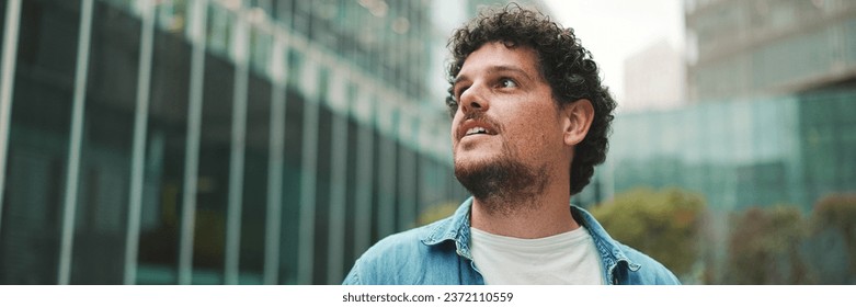 Close-up, man in denim shirt walks down the street, stops and looks around at the upper floors of modern buildings - Powered by Shutterstock