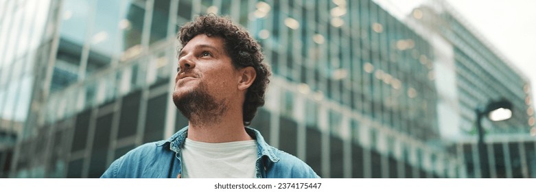 Close-up, man in denim shirt walks down the street, stops and looks around at the upper floors of modern buildings - Powered by Shutterstock