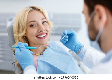 Closeup of man dentist in face mask doing treatment for patient cheerful young blonde lady smiling at camera, holding dental tools, wearing rubber gloves. Stomatology, modern dental clinic concept - Shutterstock ID 1996240583