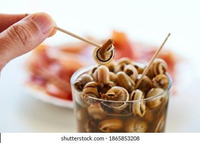 Closeup Of A Man Eating Some Caracolillos En Caldo, A Spanish Recipe Of Small Snails Cooked And Served In Broth Typical Of Andalusia, Using A Toothpick, On A Table Next To A Plate With Serrano Ham