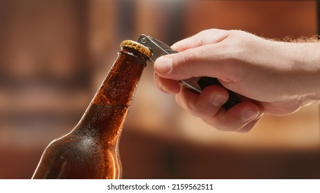 Close-up man hand opens lid fresh light beer in sweating from cold dark brown glass bottle with lid opener on blurry background. Light lager is opened with help of bottle opener.