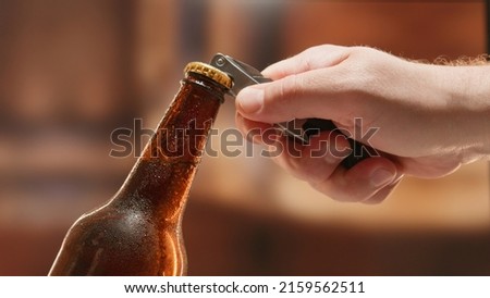 Close-up man hand opens lid fresh light beer in sweating from cold dark brown glass bottle with lid opener on blurry background. Light lager is opened with help of bottle opener.