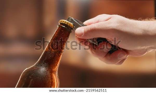 Close-up man\
hand opens lid fresh light beer in sweating from cold dark brown\
glass bottle with lid opener on blurry background. Light lager is\
opened with help of bottle\
opener.