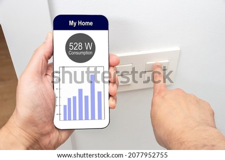 Close-up of man hand turning on the light with phone and the display of a smart electric meter with consumption information and cost per hour.