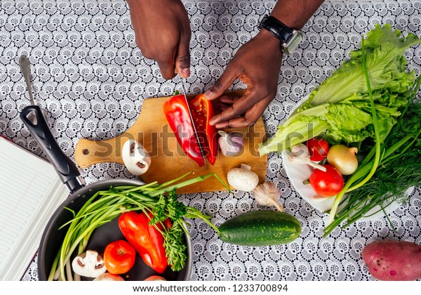 closeup of man hands african american cuts\
vegetables fry salad pepper, mushrooms, tomato in kitchen recipe\
book on the table .vegan healthy\
food