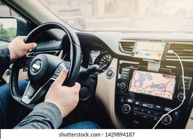 Closeup Of Man Hands Holding Steering Wheel. Person Sitting In Car Using Smartphone And Build-in Satellite Navigator GPS. Modern Gadgets In Real Life Concept. Distraction Of Attention During Driving.