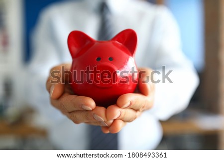 Close-up of man holding bright red piggy bank. Persons hand with container for saving money. Cash for future purchase. Money and financial well being concept