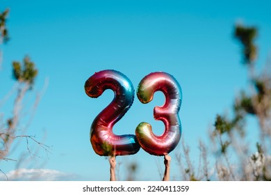 closeup of a man holding two mulitcolored number-shaped balloons forming the number 23 on the sky, standing next to some plants and trees outdoors
