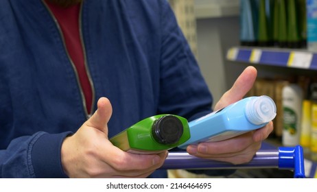 Close-up Of A Man Holding Two Shampoo Bottles In His Hands, Reading The Description And Putting One In A Shopping Cart. Selection Of Toiletries In The Store. Buying A Man In A Store.