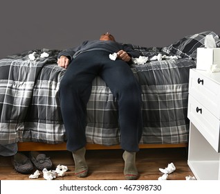 Closeup of a man laying across his bed with feet on the floor sick with the flu or COVID-19 virus unable to get up to go to work. Used tissues are strewn about the bed and floor. - Powered by Shutterstock