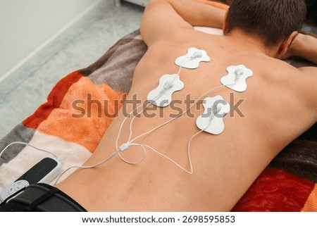 Close-up of a man lying face down with electrode pads on his back connected to an electrical stimulation device for muscle therapy and rehabilitation. Concept of physiotherapy, recovery, and wellness.