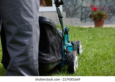 Close-up Of A Man In Overalls With A Lawn Mower Cutting Green Grass In A Modern Garden Against The Background Of A House. Lawn Mowing Machine. Professional Lawn Care Service.