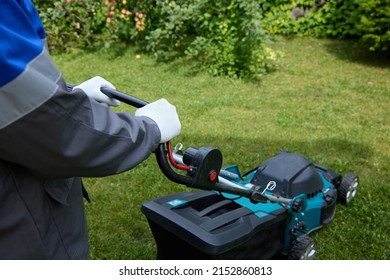 Close-up Of A Man In Overalls And White Gloves Mowing Green Grass In The Garden With A Lawn Mower. A Worker In Overalls Uses A Modern Lawnmower. Professional Lawn Care Service.