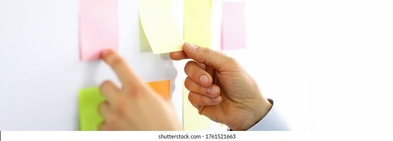 Close-up of man picking up yellow and pink lists with empty space. Persons hands and white board. Businessman in suit. Light personal cabinet. Office and business concept