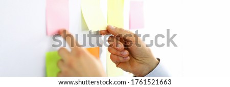 Close-up of man picking up yellow and pink lists with empty space. Persons hands and white board. Businessman in suit. Light personal cabinet. Office and business concept