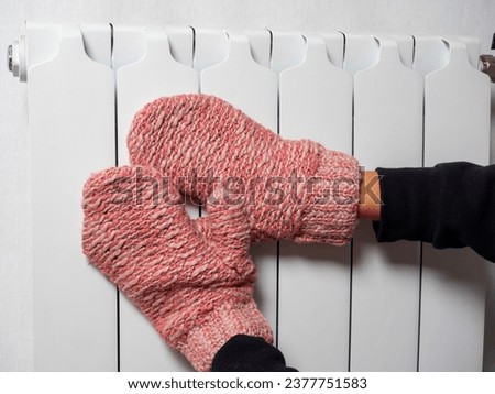 A close-up of a man puts his hands dressed in pink mittens to a white battery. The concept of the winter period, problems with central heating. Heating devices in the house, side view