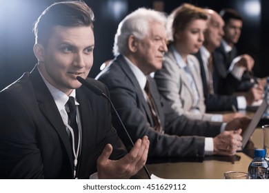 Close-up Of A Man Speaking To A Microphone During A Conference, With Its Other Participants In The Blurred Background