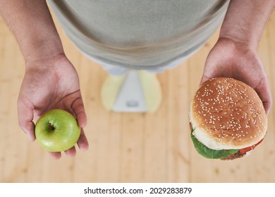 Close-up Of Man Standing On Scales Anh Holding Apple In One Hand And Burger In Other Hand To Make A Difficult Choice