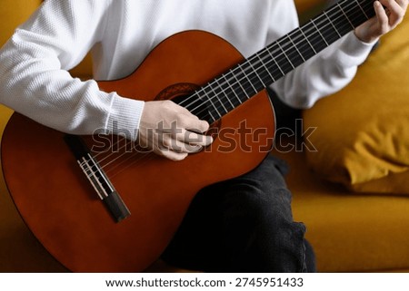 Close-up of man strumming guitar while sitting on yellow sofa indoors. Guitarist meditates while strumming strings. Concepts of creativity, hobbies, and music