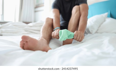 Close-up of man try to pull sock on foot, dress up to go out, sit on bed for comfort. Bright green sock, summer colour. Stylish, outfit, garment concept