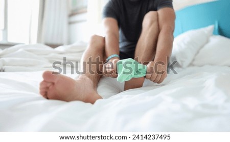 Close-up of man try to pull sock on foot, dress up to go out, sit on bed for comfort. Bright green sock, summer colour. Stylish, outfit, garment concept