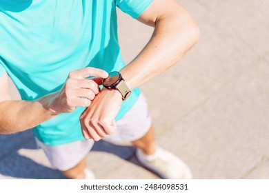 Close-up of a man in a turquoise shirt adjusting his smartwatch during a workout on a sunny day. - Powered by Shutterstock