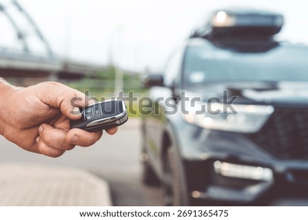 Close-up of a man unlocking a car using a key fob, with a black car parked in a car park in the background