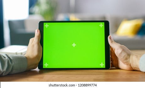 Close-up of Man Using Green Mock-up Screen Digital Tablet Computer in Landscape Mode while Sitting at His Desk. In the Background Cozy Living Room. - Powered by Shutterstock