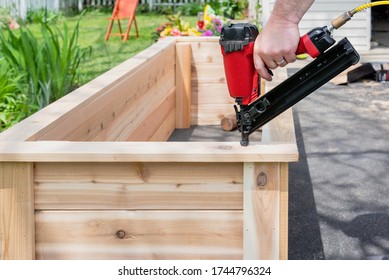 Closeup Of A Man Using A Pneumatic Nail Gun To Finish The Trim On Cedar Garden Planters With Sawdust Flying