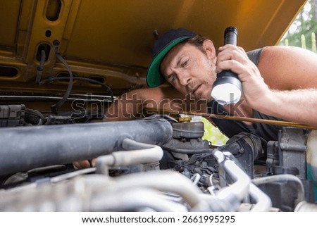 Close-up of a man wearing a cap using a flashlight to inspect a car engine. Detailed view of vehicle maintenance and mechanical work under the hood.