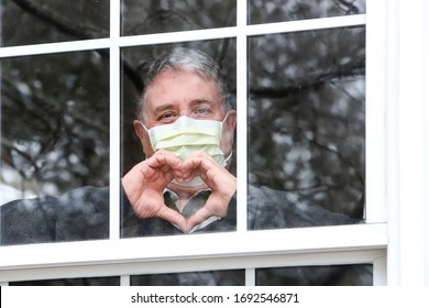 Closeup of a man wearing a face mask making heart symbol with hands at the window - Powered by Shutterstock