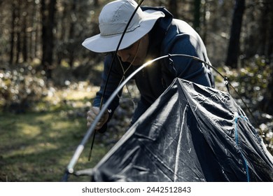 Closeup of a man wearing a hat, setting up his tent in the forest during sunset, preparing for camping adventure in the wilderness - Powered by Shutterstock