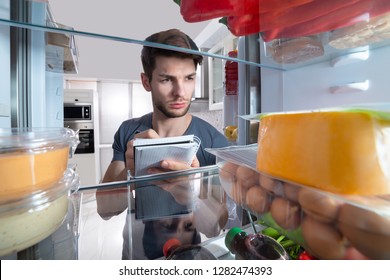 Close-up Of Man Writing On Spiral Book Near Open Refrigerator In Kitchen