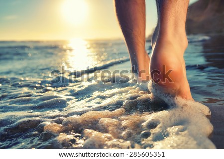 Closeup of a man's bare feet walking at a beach at sunset, with a wave's edge foaming gently beneath them, toned colors