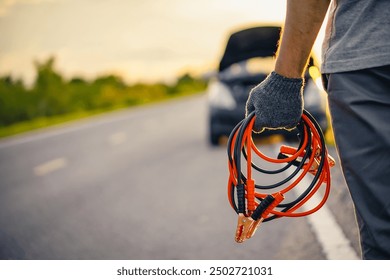 Close-up of a man's hand holding a car battery jumper cable to a broken down car on the roadside in the evening. Battery jumper cable for car battery dead