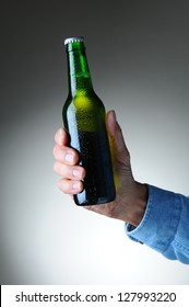 Closeup Of A Man's Hand Holding A Green Beer Bottle Over A Light To Dark Gray Background. Vertical Format. Bottle Has No Label And Is Covered With Condensation.