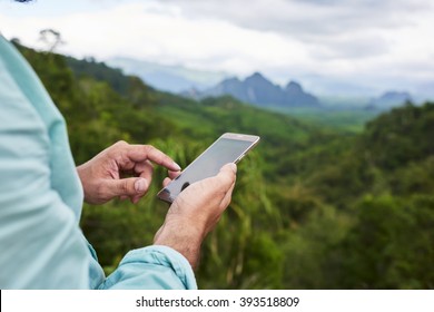 Closeup Of A Man's Hand Is Holding Mobile Phone With Copy Space Screen Against Blurred Jungle Landscape. Young Male Is Searching Information On Cell Telephone During Summer Adventure In Thailand