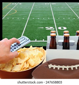 Closeup Of A Man's Hand Holding A TV Remote With A Bowl Of Chips And A Six Pack Of Beer With A Deflated Football. The TV Screen Has A Football Field With Remote Pointed At The Screen.