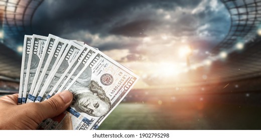 Close-up Of A Man's Hand Holding US Dollars Against The Background Of The Stadium. The Concept Of Sports Betting, Making A Profit From Betting, Gambling. American Football