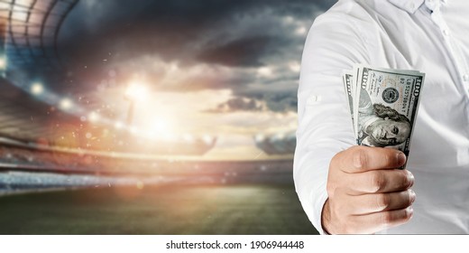 Close-up Of A Man's Hand Holding US Dollars Against The Background Of The Stadium. The Concept Of Sports Betting, Making A Profit From Betting, Gambling. American Football