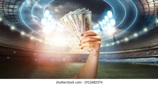 Close-up Of A Man's Hand Holding US Dollars Against The Background Of The Stadium. The Concept Of Sports Betting, Making A Profit From Betting, Gambling. American Football