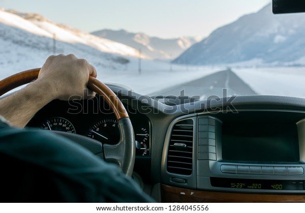 Close-up of\
a man\'s hand on the steering wheel of a car that moves on a snowy\
road among the mountains in the\
winter